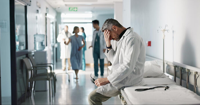 Stressed male physician sitting on hospital bed holding glasses with medical team blurred in hallway behind him.