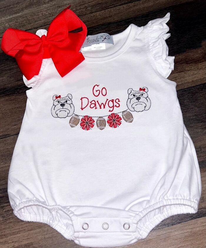 White baby onesie with red bow and "Go Dawgs" embroidery featuring bulldogs and footballs on a wooden surface, physicians' notes humor