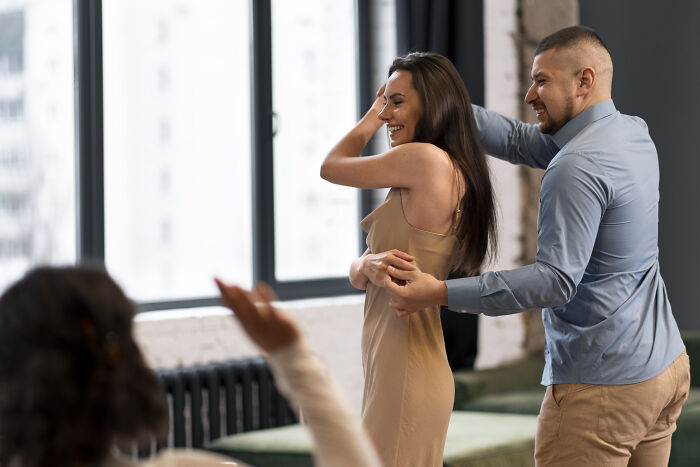 A couple laughing and dancing indoors while a third person raises a hand, capturing a funny moment related to anesthesia humor.