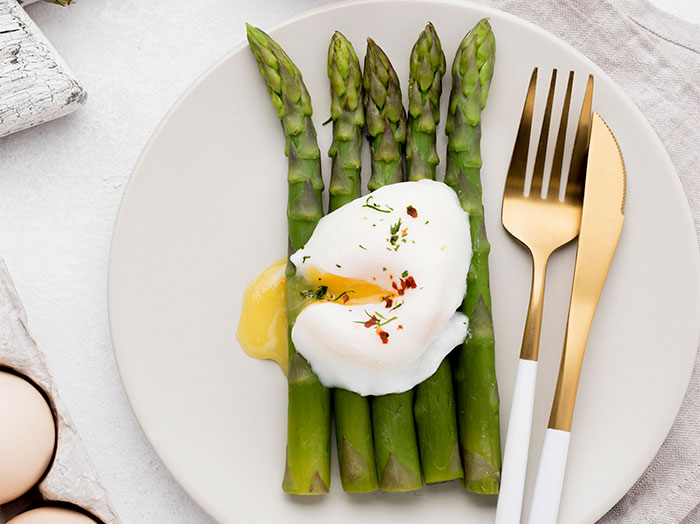 Poached egg served on fresh asparagus with seasoning and golden cutlery on a beige plate.