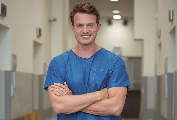 Smiling male nurse in blue scrubs standing confidently in hospital corridor, representing anesthesia and medical care.