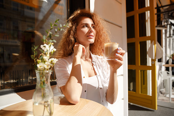 Woman enjoying coffee in a sunlit cafe, reflecting peacefully with relaxed expression, representing anesthesia humor moments.