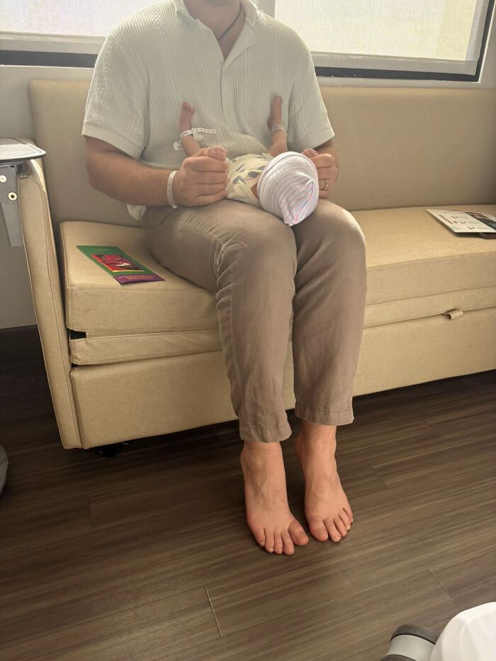 Man sitting barefoot on a beige couch holding a newborn baby, illustrating awkward body language moments from men.