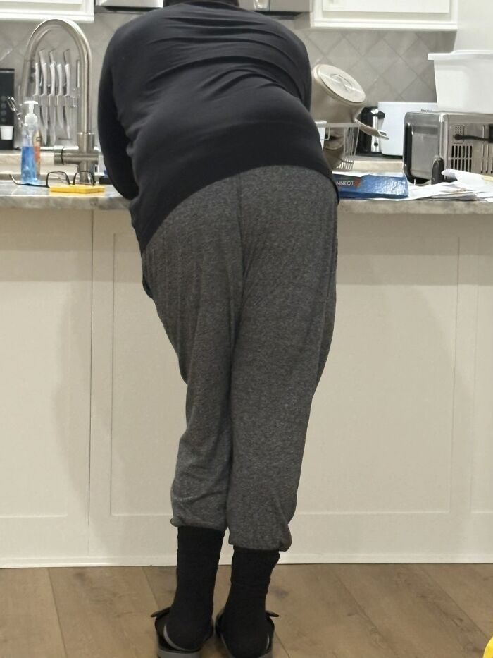 Man displaying awkward body language leaning over kitchen counter wearing casual pants and a black shirt indoors.
