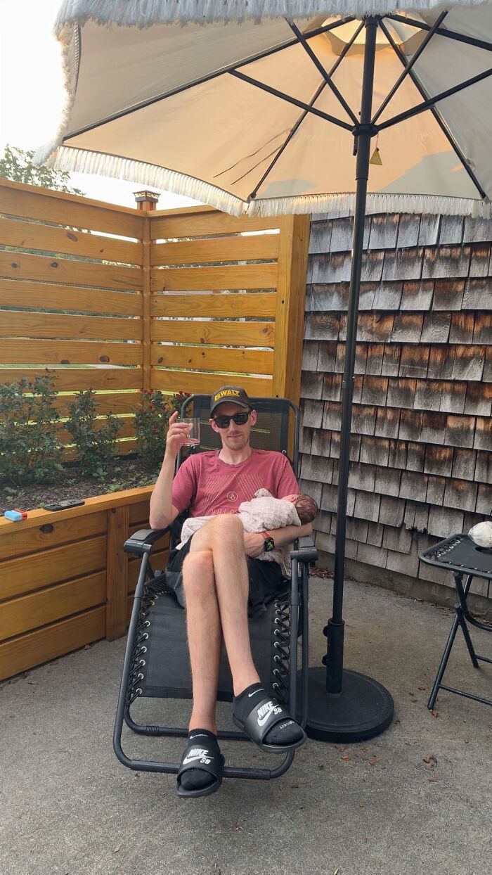 Man sitting awkwardly in outdoor chair holding baby and glass, showing hilariously awkward body language on patio.