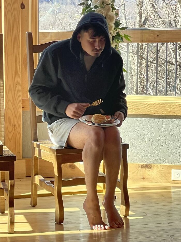 A man displaying awkward body language by sitting barefoot on a chair holding a plate of food in a wooden room.