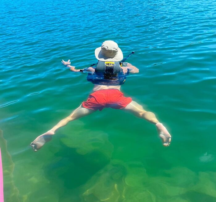 Man floating awkwardly in water wearing a sun hat and red shorts showing hilariously awkward body language.