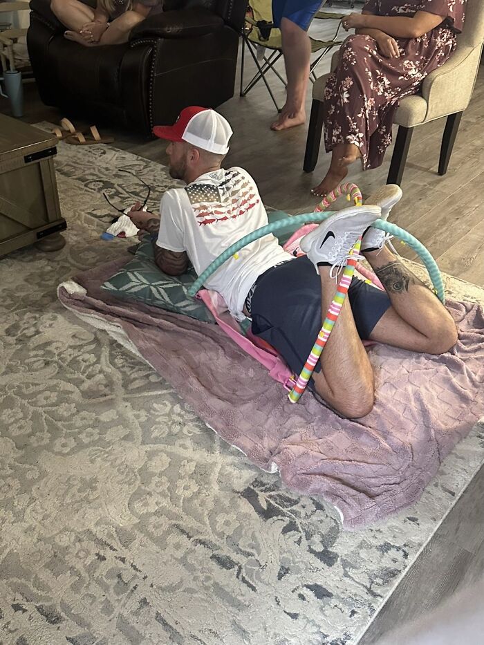 Man displaying awkward body language lying on floor wrapped in colorful hula hoops with people sitting nearby indoors.
