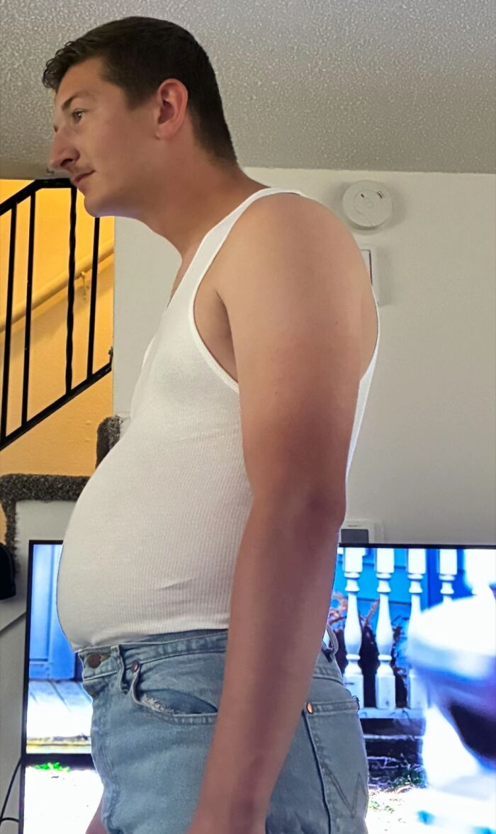 Young man in a white tank top and jeans displaying awkward body language indoors near a staircase and television.