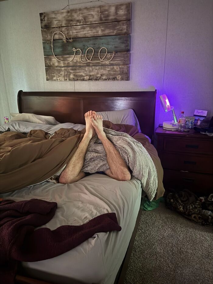 Man's awkward body language with feet tangled on bed, under blankets in a dimly lit bedroom setting.