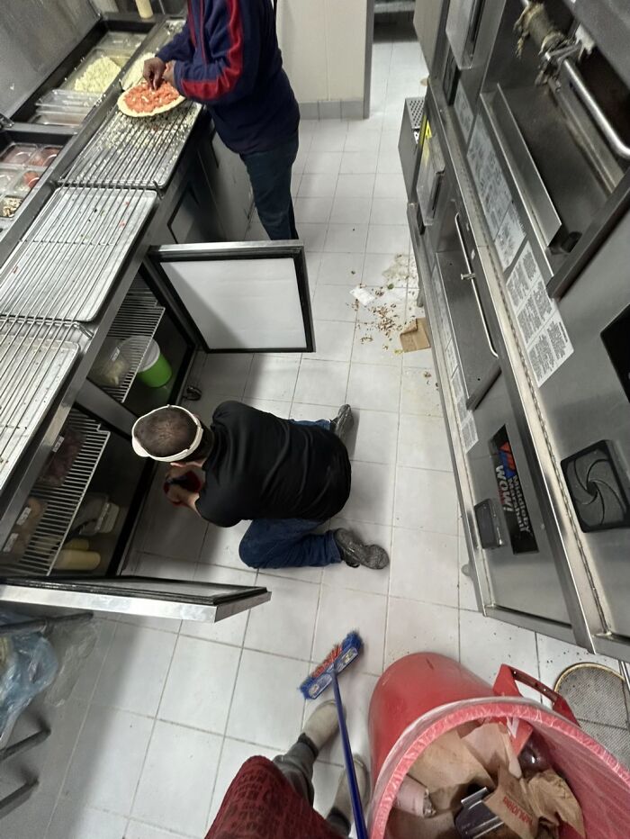 Man displaying awkward body language kneeling on kitchen floor while another prepares food in a fast-food setting.