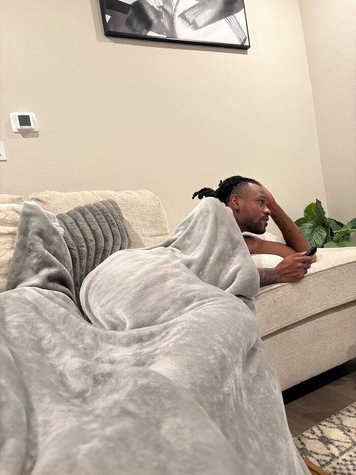 Man displaying hilariously awkward body language while lounging on a couch covered with a gray blanket indoors.