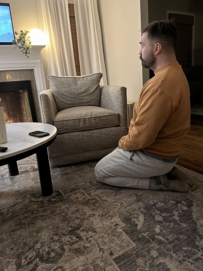 Man exhibiting awkward body language, kneeling on a carpet in a cozy living room setting near a chair and fireplace.