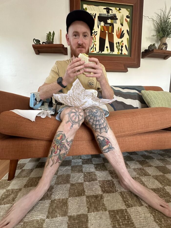 Man sitting awkwardly on a couch with legs spread wide, showing tattoos and holding a sandwich, illustrating awkward body language.