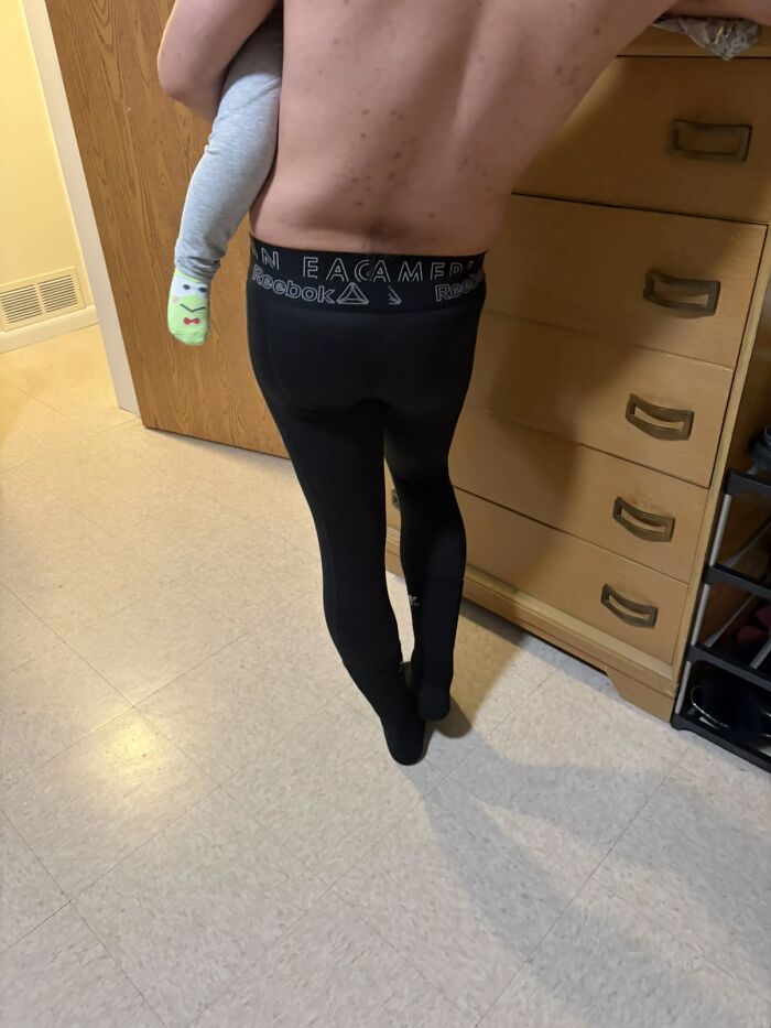 Shirtless man wearing black leggings awkwardly holding a child indoors, illustrating men’s hilariously awkward body language moments.