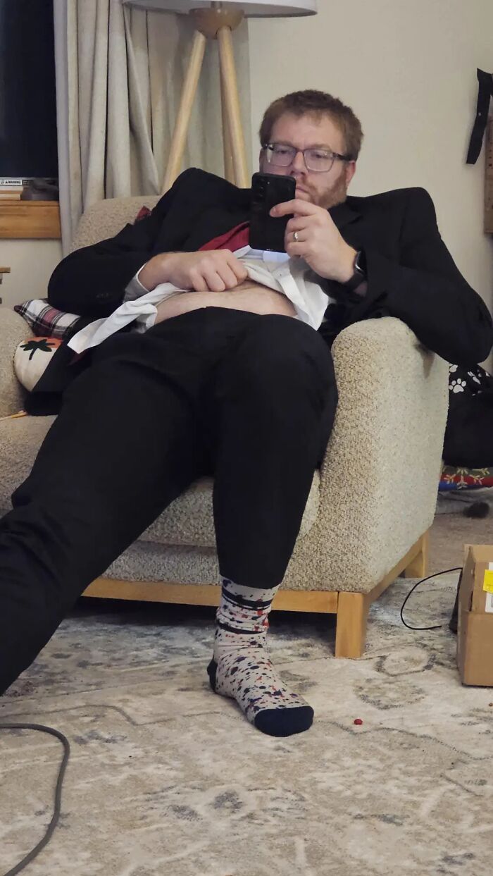 Man displaying hilariously awkward body language slouching in chair with shirt lifted and looking at phone.