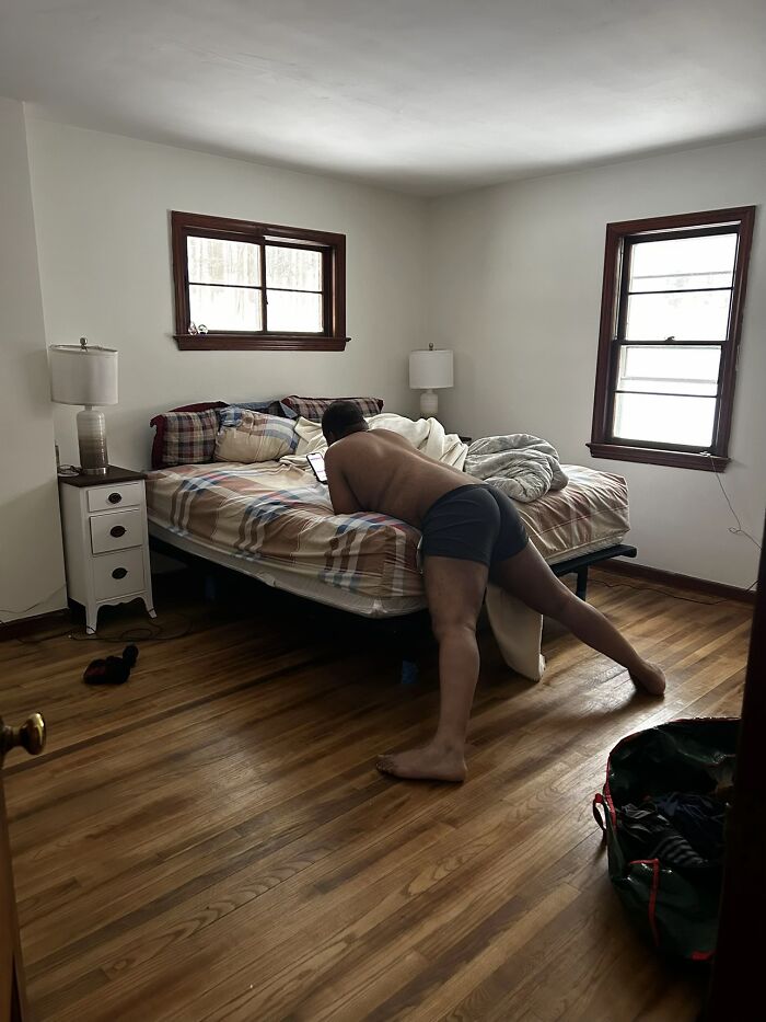 Man in dark shorts leaning awkwardly on a bed in a simple bedroom, illustrating hilariously awkward body language.