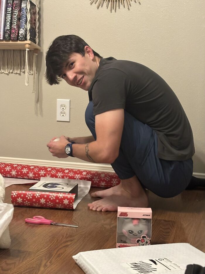 Young man squatting awkwardly on the floor while wrapping gifts, showing humorous men’s body language moments.