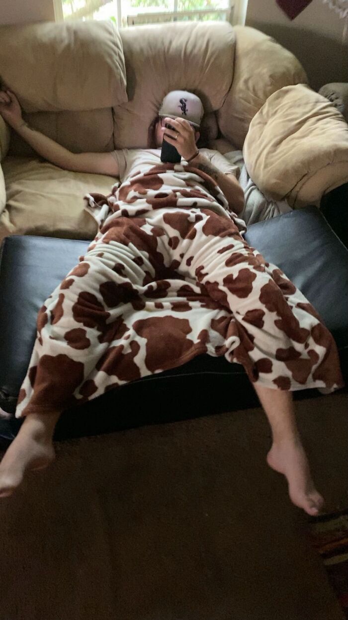 Man lying awkwardly on couch in cow-print blanket pants, looking at phone, showcasing hilariously awkward body language.