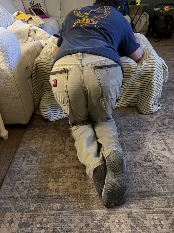 Man kneeling awkwardly on a patterned rug leaning over a couch, displaying awkward body language indoors.