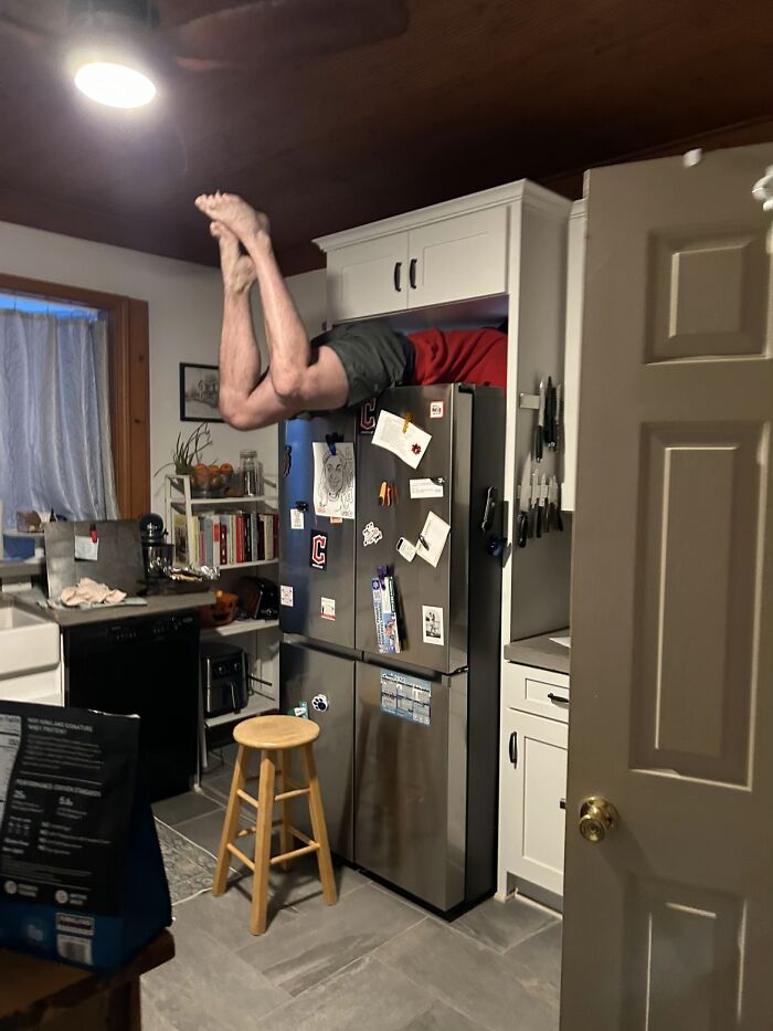 Man displaying hilariously awkward body language by hanging upside down half inside a kitchen cabinet above a refrigerator.
