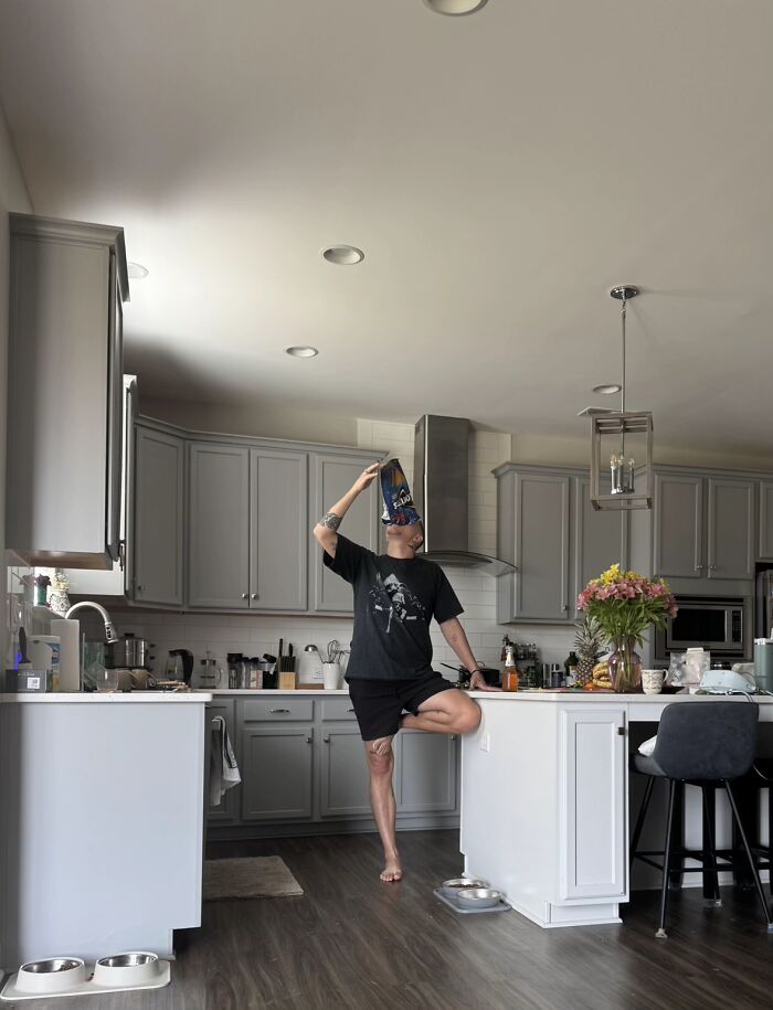 Man displaying awkward body language standing on one leg in kitchen while drinking from large snack bag, illustrating men’s body language.