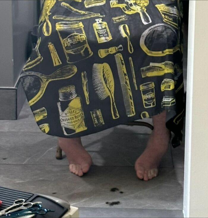 Bare feet awkwardly positioned under a barber's cape with grooming tools pattern, showing men’s awkward body language.