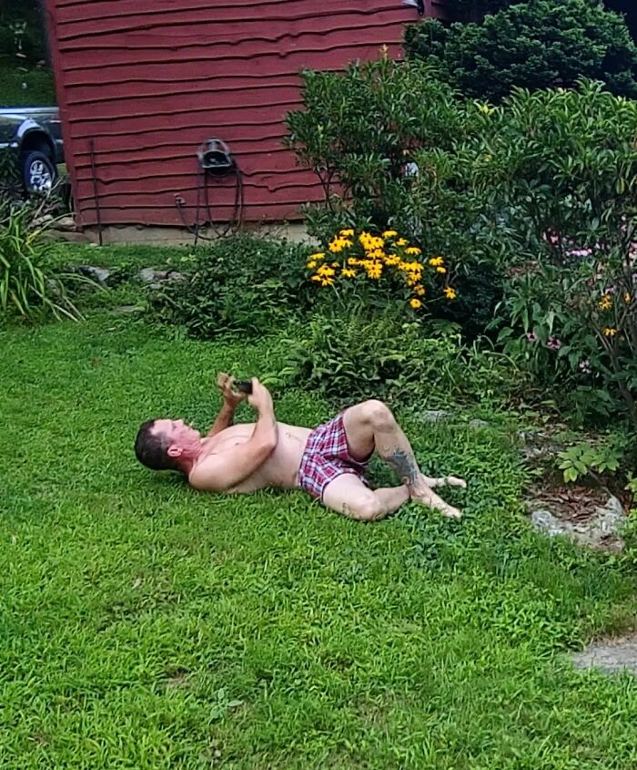 Man lying awkwardly on grass in plaid shorts, displaying hilariously awkward body language near garden flowers.