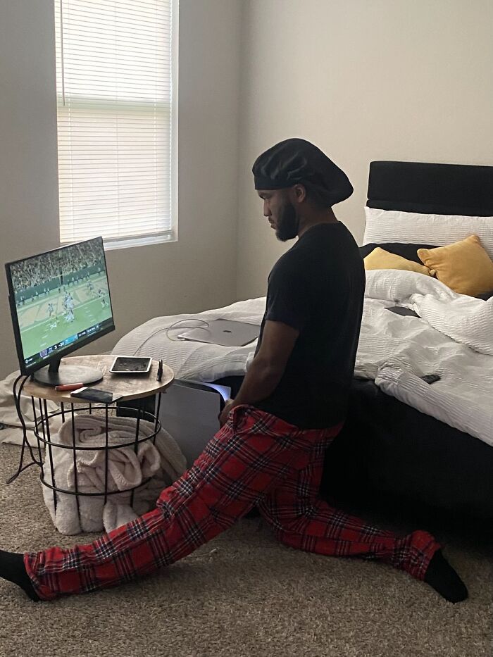 Man in red plaid pants awkwardly stretching in a bedroom while watching football, showing awkward body language.