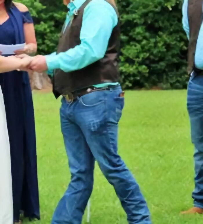Man with awkward body language wearing blue jeans and a vest outdoors during a casual social interaction.