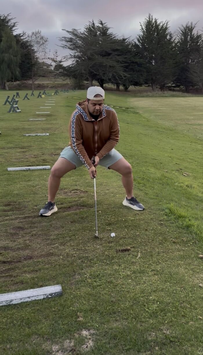 Man exhibiting hilariously awkward body language while preparing to hit a golf ball on a grassy course.
