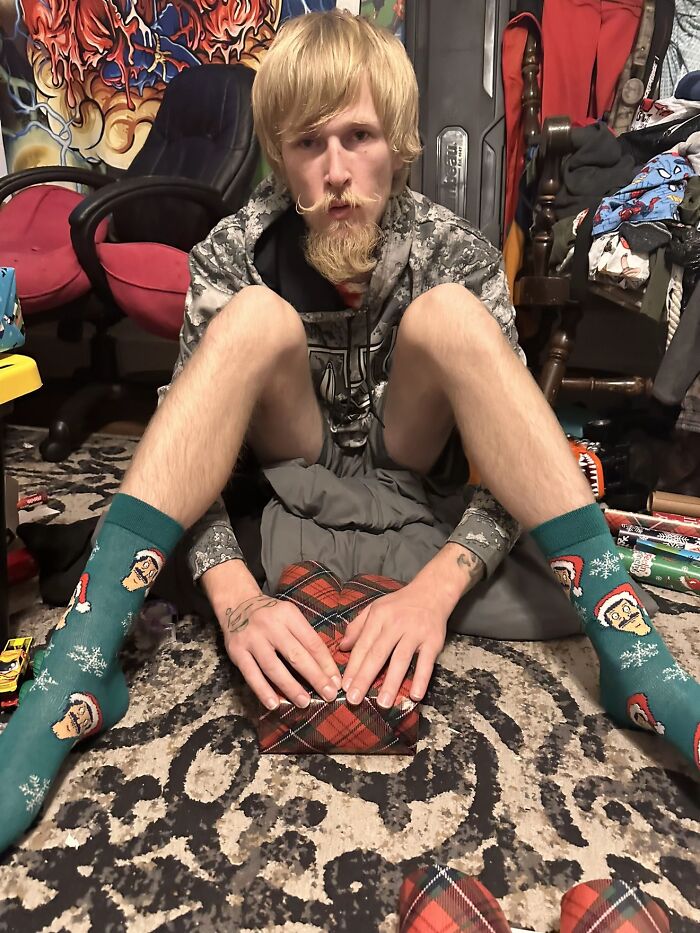 Young man displaying hilariously awkward body language while sitting on the floor wrapping gifts in a cluttered room.