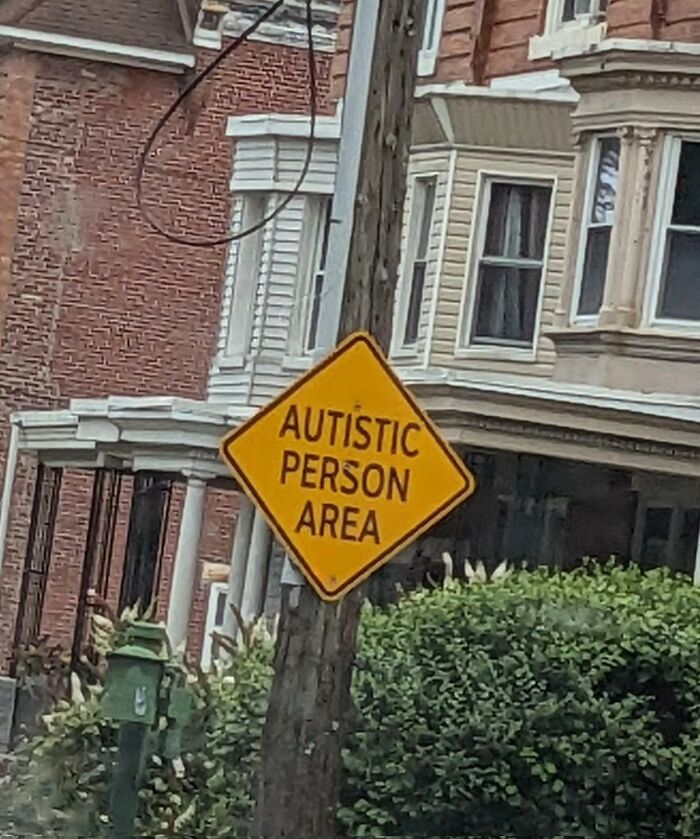 Yellow street sign reading autistic person area posted on a wooden pole near residential buildings and greenery in a neighborhood.