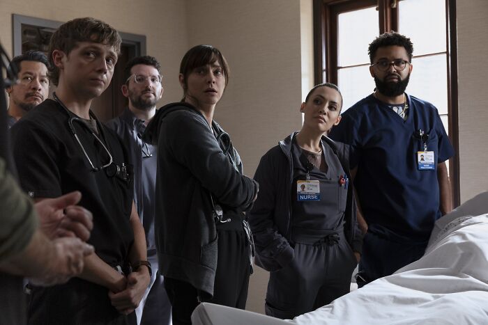 Cast members from The Pitt Season 2 Episode 6 standing in a hospital room, dressed as medical professionals, observing a patient. - 8