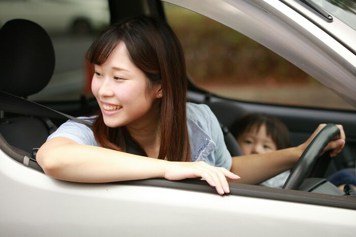 Young woman smiling inside a car with a child in the driver’s seat, illustrating girls and problematic childhood norms.