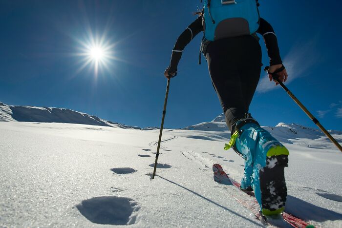 Skier with bright blue boots trekking across snowy terrain under a clear blue sky in Olympic sports winter setting.