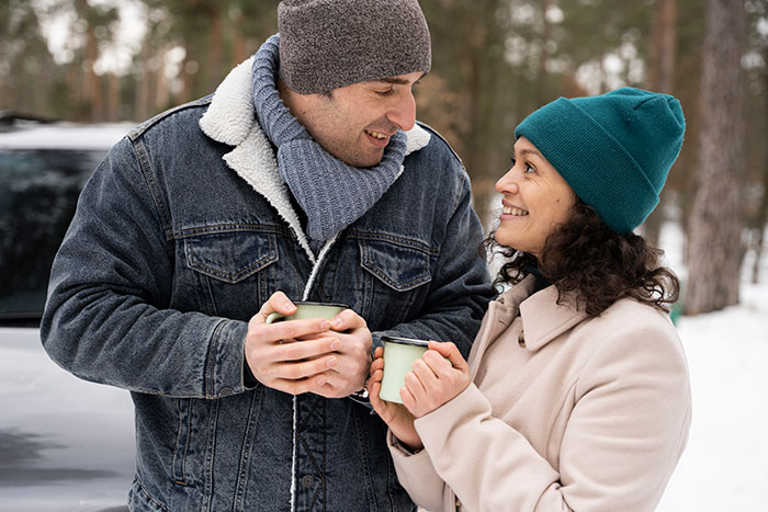 Couple outdoors in winter holding mugs, the boyfriend looking suspiciously at his girlfriend’s second phone. Couple outdoors in winter holding mugs, the boyfriend looking suspiciously at his girlfriend’s second phone.