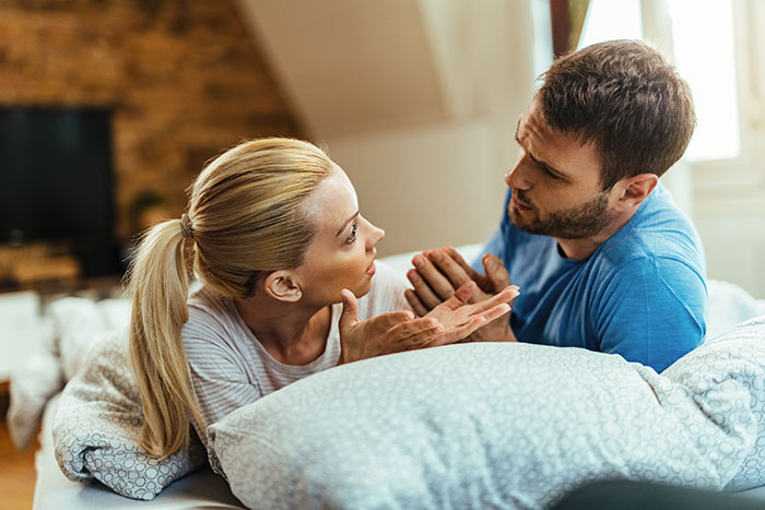 Couple lying on bed, woman appearing frustrated and man looking defensive, depicting suspicious boyfriend with second phone. Couple lying on bed, woman appearing frustrated and man looking defensive, depicting suspicious boyfriend with second phone.