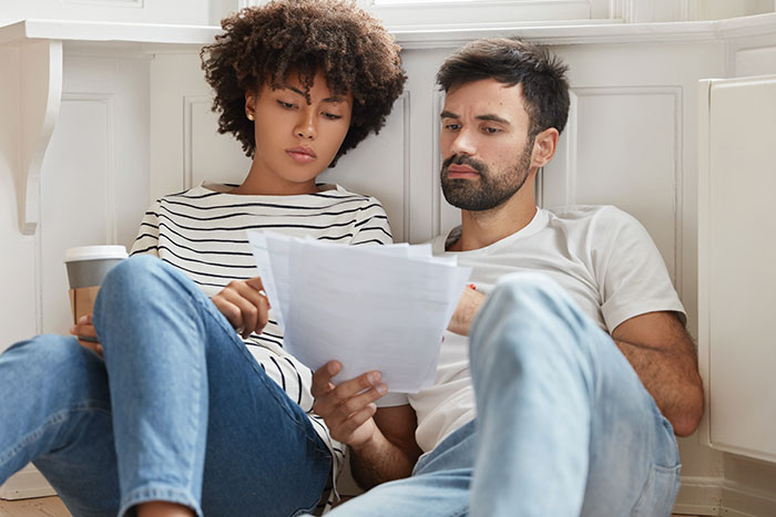 Couple sitting on floor, reviewing documents with serious expressions, reflecting concerns about GF quitting job secretly.