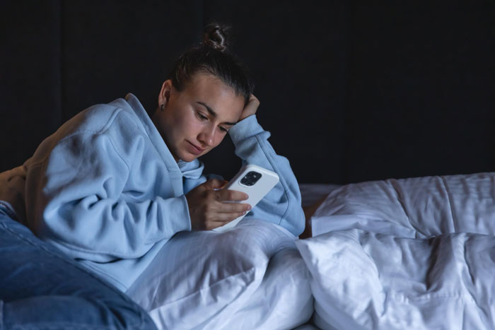 Young woman in a blue hoodie lying on bed, looking at phone with a thoughtful expression, showing ghoster relationship messages.