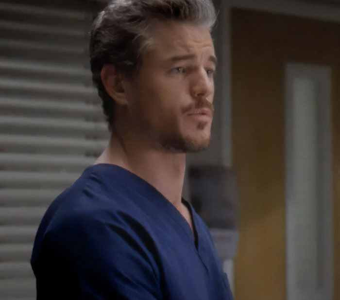 Eric Dane in character on Grey's Anatomy, wearing blue scrubs and looking contemplative in a hospital setting.