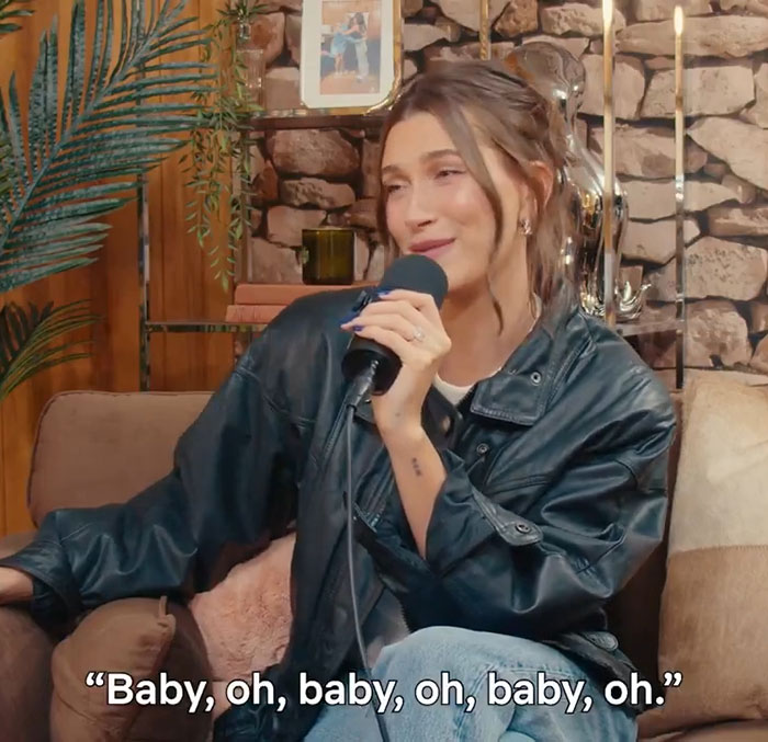 Hailey Bieber speaking into a microphone, seated casually in a cozy room with plants and decorative stone wall background.