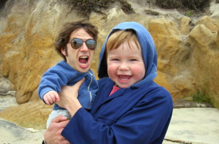 Toddler with adult face wearing blue hood and jacket holding an angry adult with a small body in an outdoor rocky area, photoshop effect.