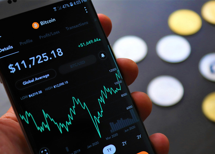 Hand holding smartphone displaying Bitcoin price chart and exchange data with cryptocurrency coins blurred in background