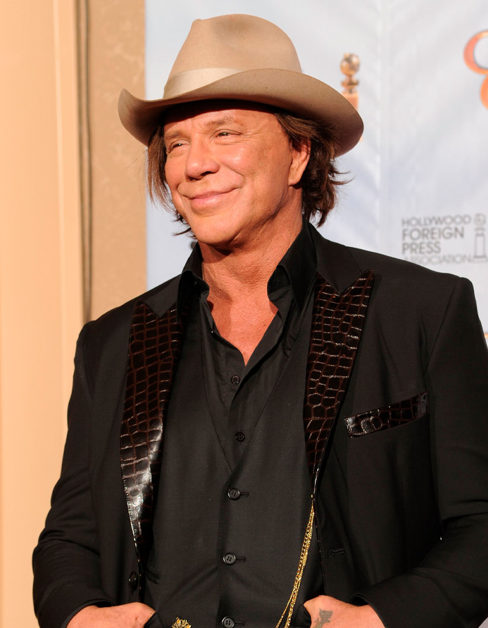 Mickey Rourke smiling in a beige hat and black suit with crocodile leather details at a formal event.