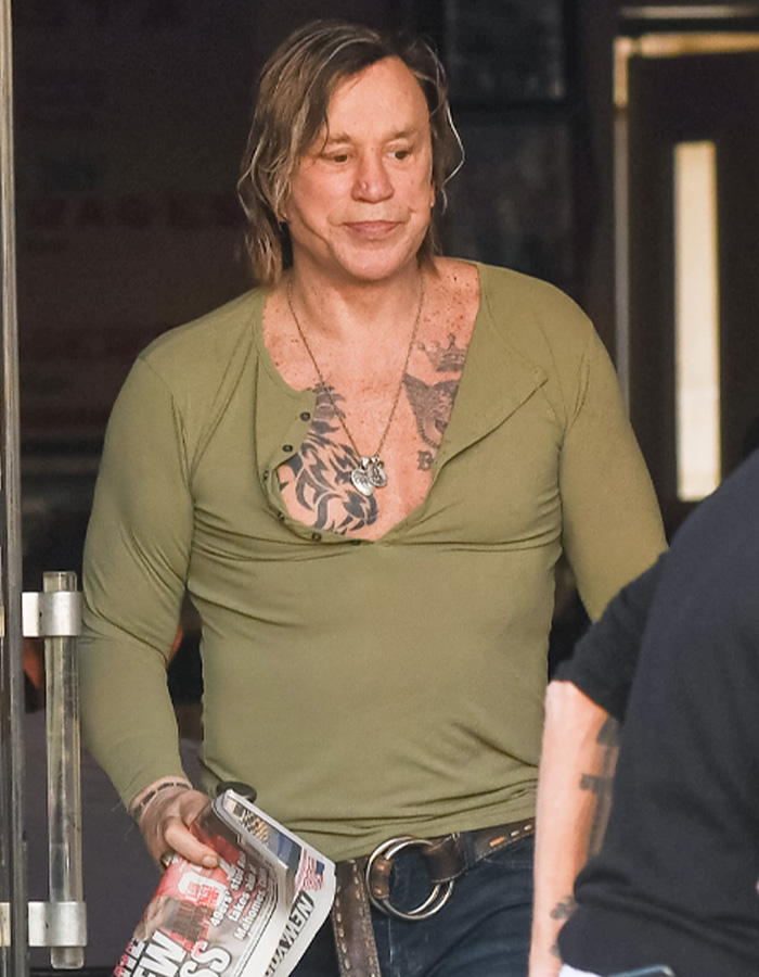 Mickey Rourke wearing a green shirt showing tattoos, holding a newspaper, and walking outdoors looking casual.