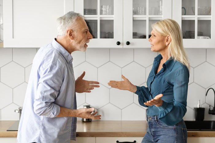 Middle-aged woman and older man arguing in modern kitchen, reflecting conflict between cheating husband's mom and in-laws.