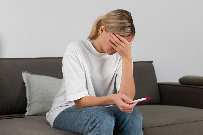 Woman sitting on couch, distressed and holding a pregnancy test, reflecting on husband best friend cheating situation.