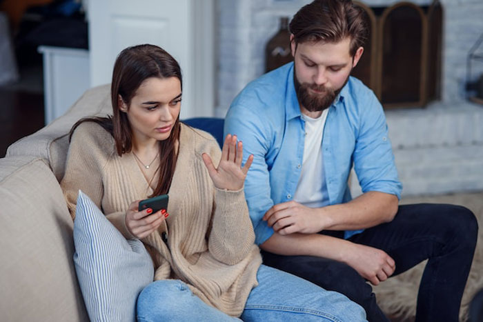 Woman upset holding phone while man sits beside her showing tension in husband best friend cheating situation at home