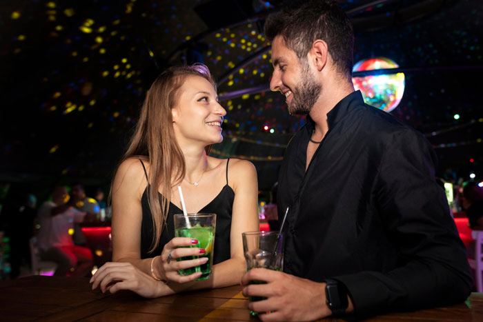 Woman playing wing woman for man-crazy friend, smiling and holding drinks in a dimly lit bar with colorful lights.